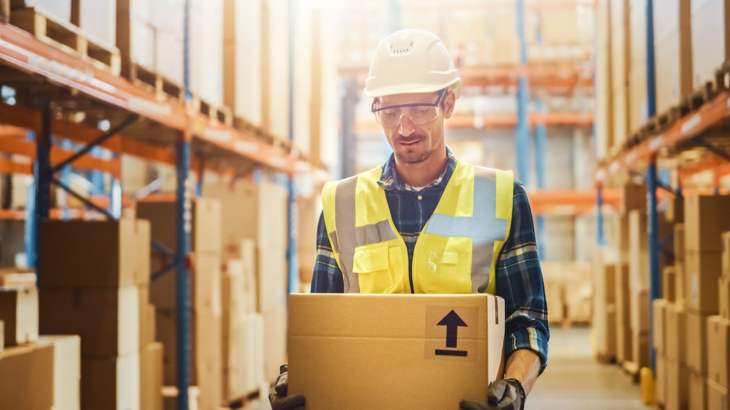 handsome-male-worker-wearing-hard-hat-holding-cardboard-box-walking-through-retail-warehouse-full-of-shelves-with-goods-working-in-logistics-and-distribution-center-front-shot